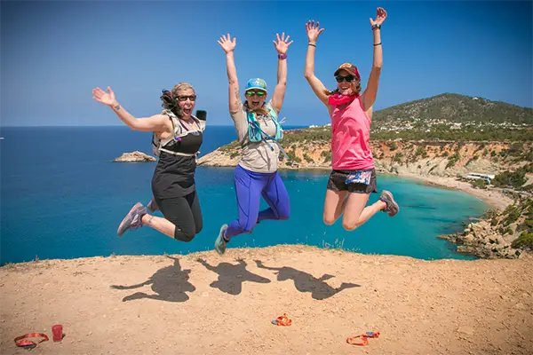 Race2Adventure Greece 2028 global vacation adventure three smiling women runners jump into the air atop a sunny cliff with azure waters and beaches beyond. Run, seek, adventure!