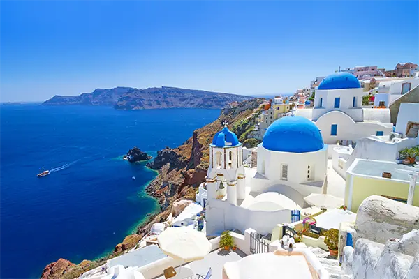 Race2Adventure Greece 2028 global vacation adventure aerial view of the greek island Santorini featuring white buildings with cobalt domed rooftops surrounded by turquoise waters on a sunny day. Run, seek, adventure!