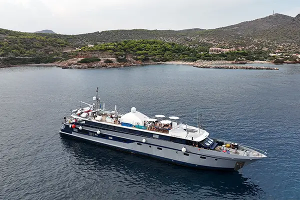 Race2Adventure Greece 2028 global vacation adventure aerial view of a white ship anchored in azure waters off a Greek island on a sunny day. Run, seek, adventure!